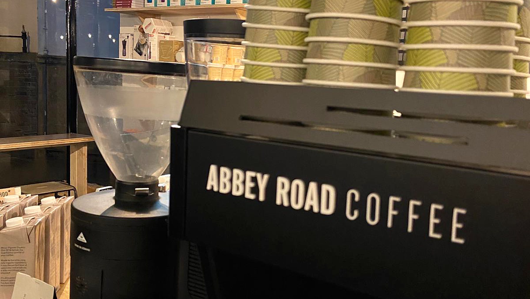 Close-up of the black espresso machine with Abbey Road Coffee lettered across it, a coffee grinder beside it and stacks of paper cups on top