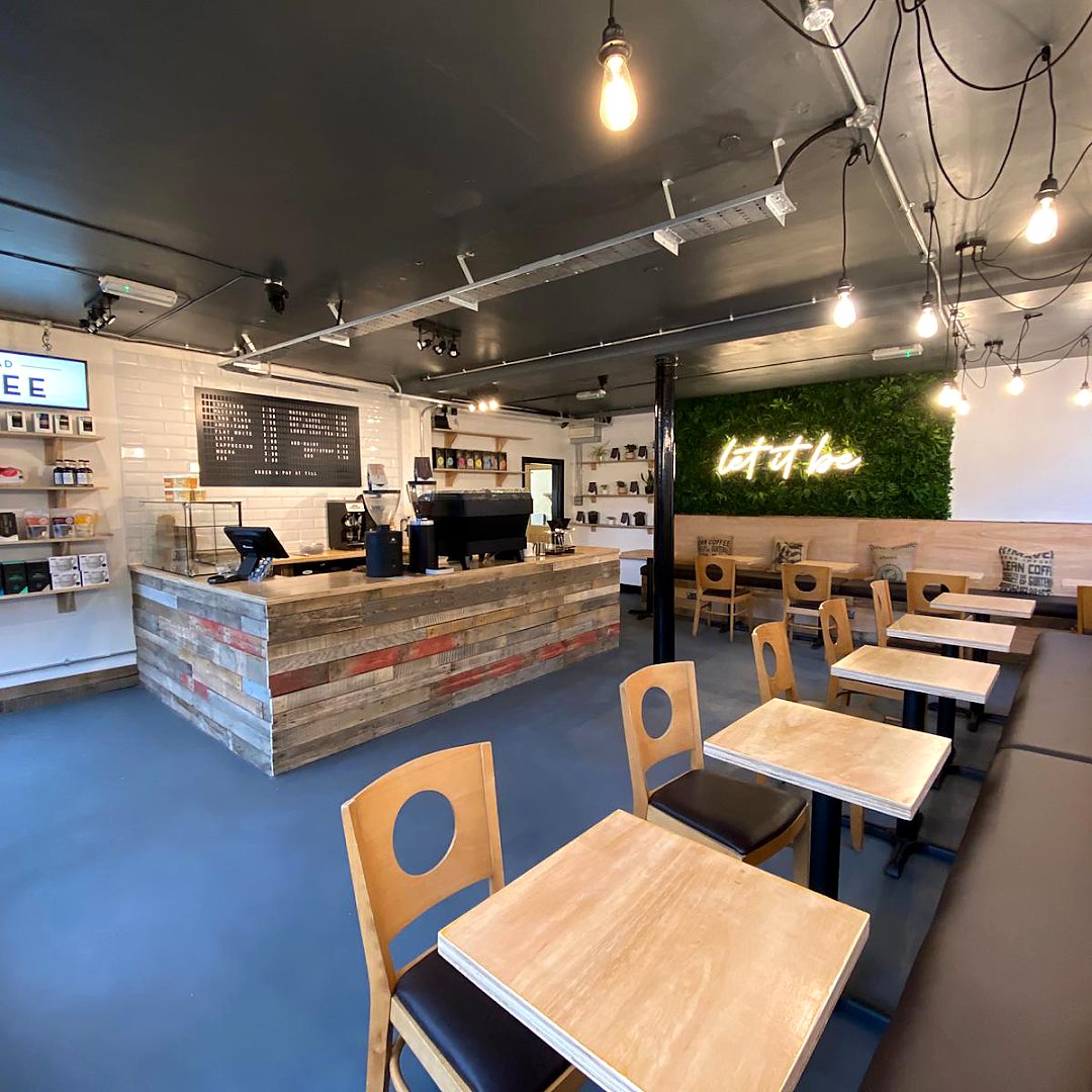 Inside the café: light wooden tables and chairs, a counter clad in reclaimed planks, filament bulbs hanging from the ceiling and a green plant wall with a neon sign