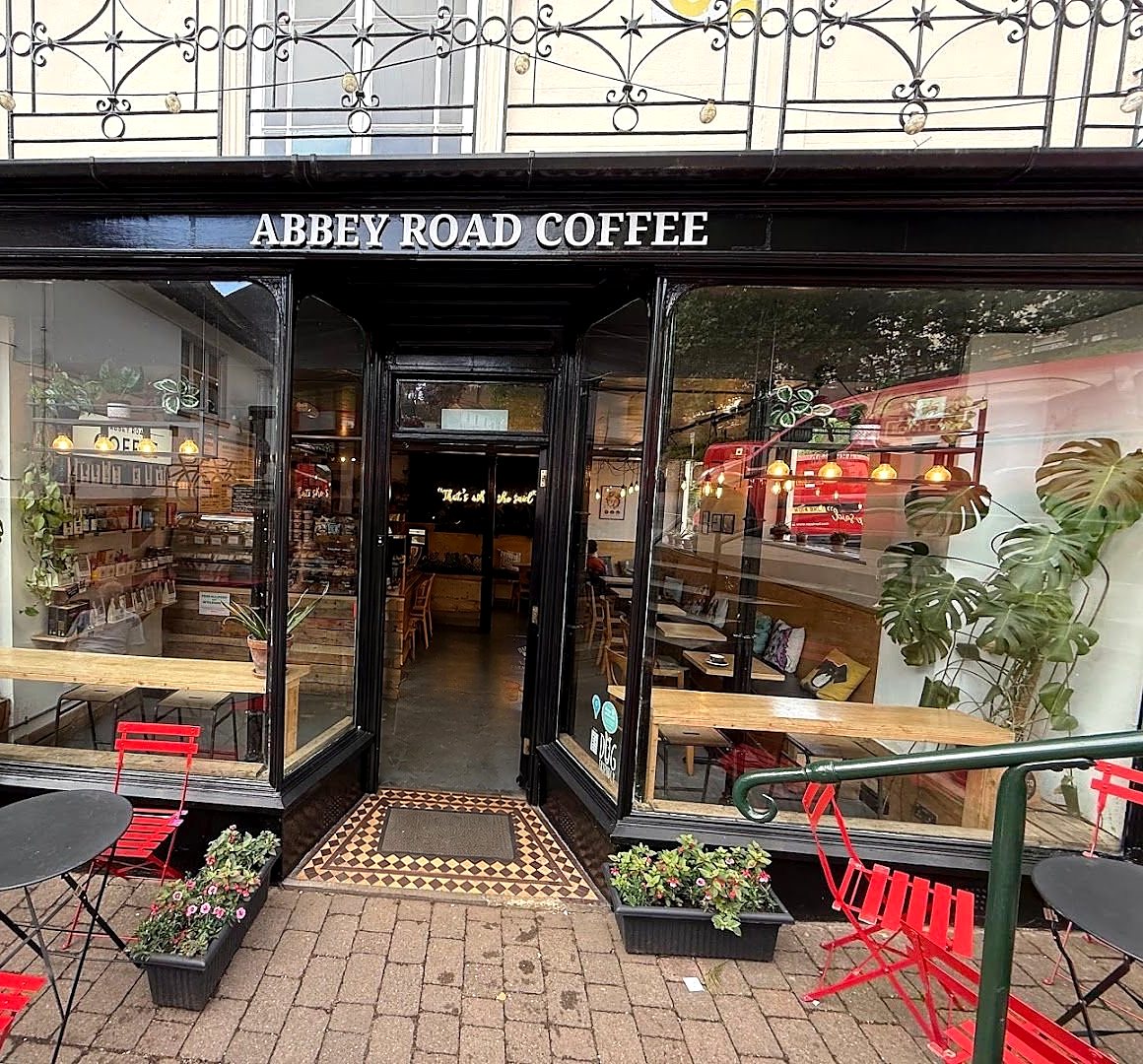 The Abbey Road Coffee shopfront: a black frontage with the name in white letters, big windows full of plants, an open door onto a tiled step, and red folding chairs at small black tables on the pavement